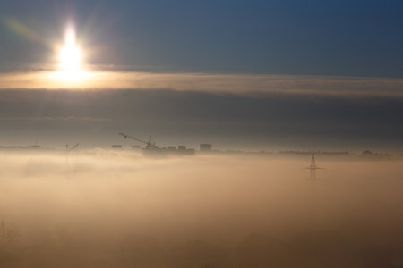 Sunrise in the city, foggy morning, silhouettes of multi-storey buildings, construction cranes and high-voltage poles in the fog. city landscape at dawn. Thick low fog and bright rising sun.の写真素材