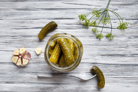 Pickled cucumbers in an open jar, gherkin on a fork and garlic on a light wooden background, top view. Homemade canned cucumbers.の写真素材