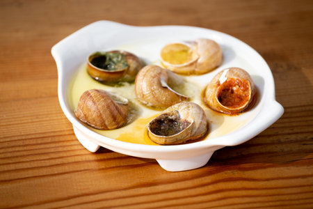 Cooked snails with garlic, tomato, spicy sauce, herbs, white wine on a white dish on a wooden background, gourmet french cuisine, Escargots de Bourgogne.の写真素材