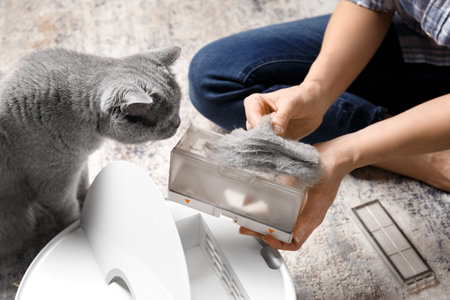 The container of the vacuum cleaner robot is in female hands, the dust collector of the robot vacuum cleaner is full of wool, the cat sits nearby and looks at the container.の写真素材