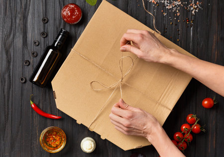 Pizza in a closed cardboard box, sauces, fresh vegetables on a dark wooden background, female hands unpack pizza, top view. food delivery.の写真素材
