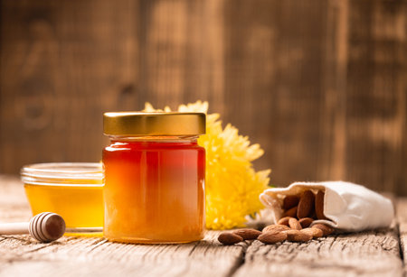 Honey in a closed glass jar and a bowl, sticks for honey and scattered almonds on a wooden background. Composition of honey jar, dipper, nuts in a bag close-up. Healthy foods.の写真素材