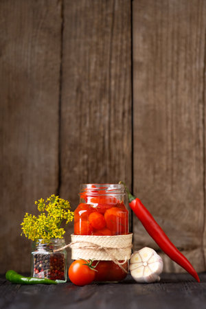 Pickled cherry tomatoes in an open glass jar, chili peppers, garlic, spices and herbs for marinade on a dark wooden background. home preservation.の写真素材