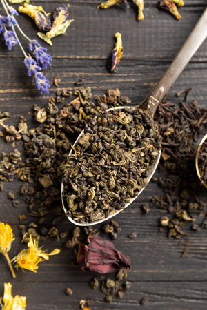 Composition of dry black and green tea in vintage spoons and dry hibiscus, calendula, lavender flowers on a dark wooden background top view close-up. Different types of dry tea on a dark background.の写真素材