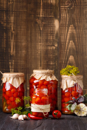 Canned cherry tomatoes in closed and open jars, garlic, hot peppers, basil on wooden, space for text. Pickled tomatoes, spices and herbs. Ingredients for Preserving Vegetablesの写真素材