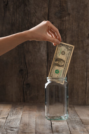 A hand puts one paper dollar in a glass jar on a wooden background. The concept of savings, storage, safety, saving money, distrust of banks.の写真素材