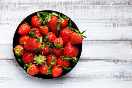 Fresh organic strawberries in a black round plate on a light wooden background, top view, with space for text.の写真素材