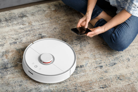 A girl sits on the floor in the living room and controls a robotic vacuum cleaner using a smartphone. smart home. The concept of smart home appliances, housework and technology.の写真素材