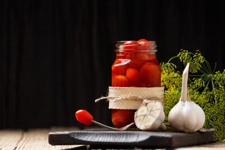 Pickled cherry tomatoes in an open jar on a dark wooden board, one tomato on a vintage fork over a jar, dark background, close-up.の写真素材