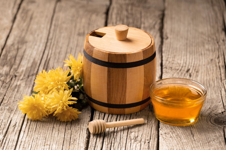 Honey in a small wooden barrel and a transparent glass bowl, a wooden dipper, yellow flowers on an aged wooden background, close-up.の写真素材