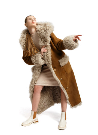 A young stylish girl in a luxurious long brown sheepskin coat with a fur hood and cuffs poses beautifully on a white background. Fashionable, stylish, comfortable outerwear.の写真素材