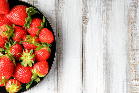 Fresh organic strawberries in a black round plate on a light wooden background, top view, with space for text.の写真素材