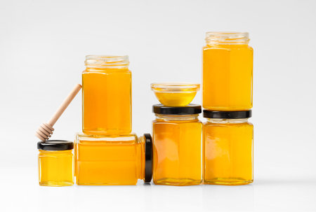Conceptual composition of glass jars in the form of honeycombs with honey and wooden dipper on a white background. Creative composition of closed and open honey jars one on top of the other.の写真素材