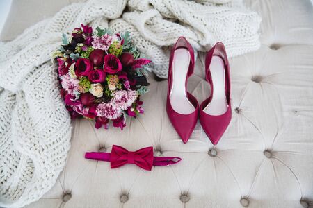 Bridal bouquet with red peonies, bow tie and red high-heeled shoes on a white pouffe, next to a knitted white blanketの写真素材