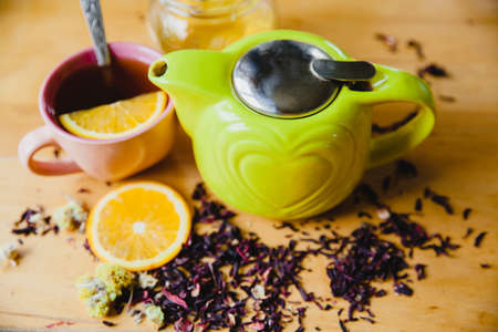 Green teapot with heart on a wooden table. Next to a slice of orange, full cup of tea, honey jar and scattered tea leaves and herbs.の写真素材