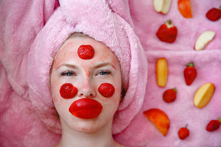 A young girl with a pink towel on her head lies with a mask of strawberries on her face. Nearby pieces of fruitの写真素材