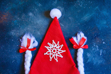 Santa Claus hat, braids with red bows and a knitted white snowflake on a blue grunge background. New...
