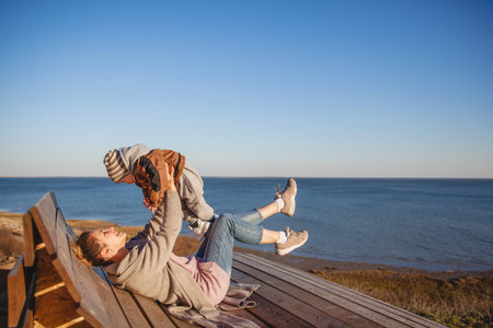 A young woman holds her baby at arms length and laughs as she lies on a bench on the seaside in...
