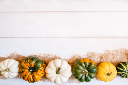 A row of multicolored small pumpkins on burlap on a white wooden background. Flat layout with space...
