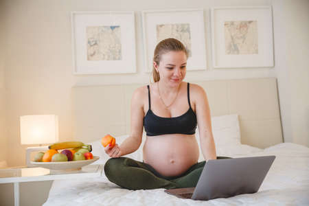 A young blonde pregnant woman is sitting on the bed and eating fruit while looking at her laptopの写真素材