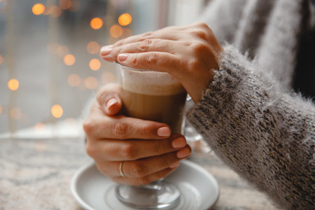 A woman in a warm jacket is sitting at a table by the window and warms her hands on coffee on the...