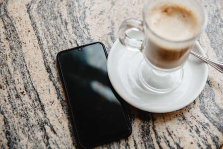 A glass of coffee on a saucer on the table and a black smartphone on the marble table. High quality photoの写真素材