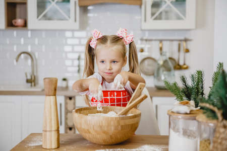 Four-year-old blonde girl in a white t-shirt and red apron kneads dough at home in the kitchen at a wooden tableの写真素材
