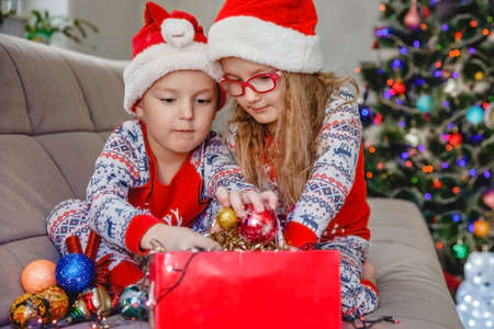 Little brother and sister in Santa hats on the sofa against the the Christmas tree take out decorations from the boxの写真素材