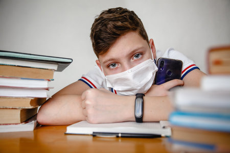Portrait of a teenager at home in a mask behind textbooks. In the hands of a smartphone.