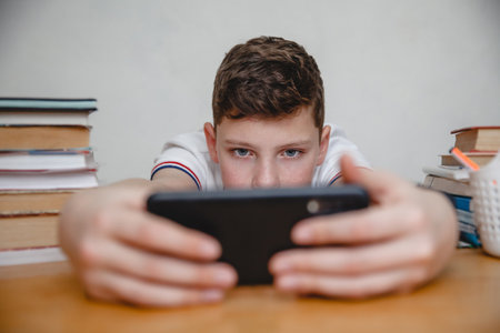 A teen looks into a black smartphone holding it at arm's length at home at a table among textbooks. High quality photoの写真素材