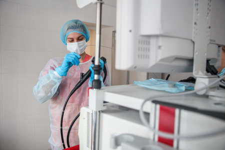 A female endoscopist surgeon in a protective suit, cap, mask and gloves holds an endoscope in front of a monitor. High quality photoの写真素材