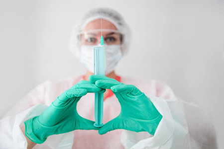 A medical worker in a white robe, white cap and green gloves holds a syringe with a vaccine....
