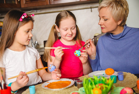 Blonde mom shows her daughters how to decorate Easter eggs at home in the kitchen. High quality...