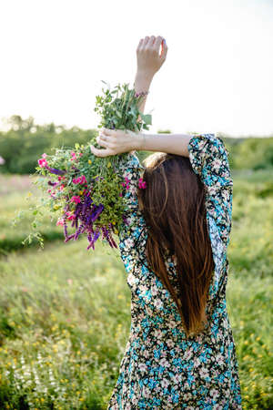 A young woman with long hair in a colorful short dress holds a bouquet of wild flowers in her raised hands while standing with her back to the cameraの写真素材