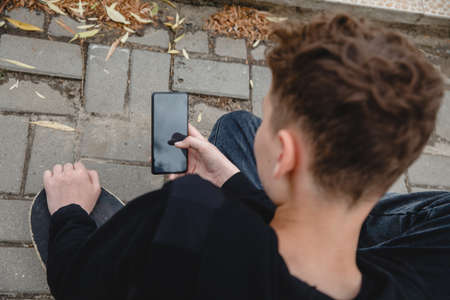 A curly-haired teen of European appearance in black hoodie sitting on an alley on a skateboard and talking on the phoneの写真素材