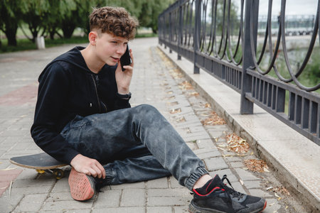 A curly-haired teen of European appearance in black hoodie sitting on an alley on a skateboard and talking on the phoneの写真素材