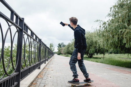 A curly-haired teenager of European appearance in a black hoodie standing on a skateboard in an...