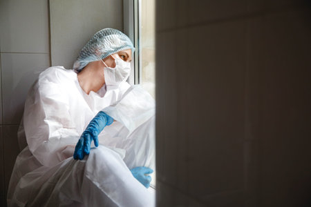 a young female doctor in a protective suit, cap, mask and gloves is sitting wearily near the window...