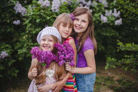 Three smiling European girls stand hugging in a summer garden with lilac flowers in their handsの写真素材