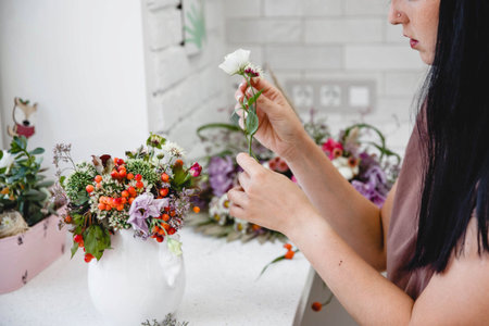 A brunette European woman florist with long hair makes a bouquet in a white vase at a white table in...