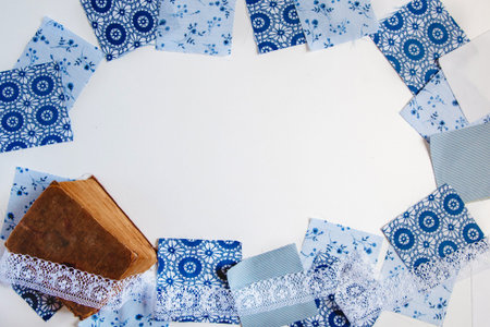 Square patches of blue printed fabric, old shebby book and white lace on a white background. Textile...