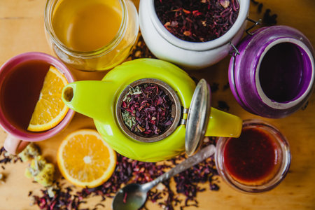 Open green teapot with tea leaves and herbs. Around it honey, leaf tea and jam in jars. Also next to...
