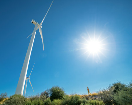 Huge wind turbine and tiny human silhouette on top of a hill against blue sky and shining sun, with...