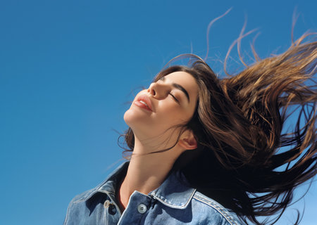 Young woman with long dark hair blowing in the wind against a blue skyの素材