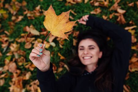 Beautiful woman laying on a green grass in a park at the grass. Sporty young brunette female athlete. Fitness woman take a rest at fall park. Surrounded by autumn leaves on green lawn. Focus on leaf.の写真素材