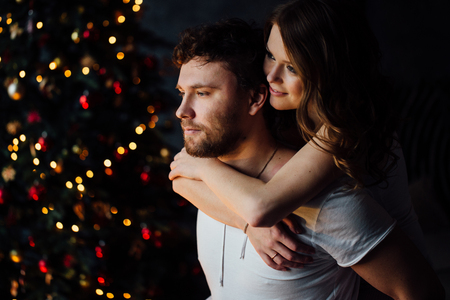 Beautiful young couple in pajamas on nicely decorated Christmas tree background. Girl hugs a guy from behind.の写真素材