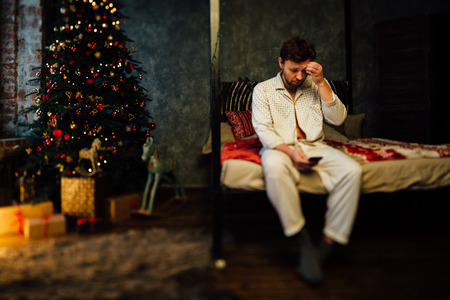 Confused man wearing pajamas sitting on the bed and looking at the phone, scratching his forehead christmas tree background.の写真素材