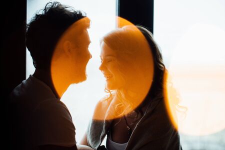 Happy couple of lovers in pajamas sit on the windowsill. Silhouette Close up. Garland lights in the foreground bokeh. Soft focus atmosphere.の写真素材