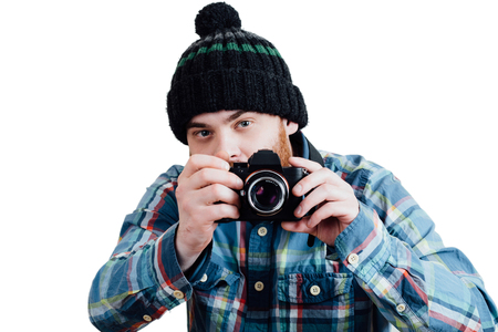 Portrait of a red-bearded, balding male brutal. White isolated background. A man in a blue plaid shirt with knitted cap. Holding camera in the hands.  Ready to shootの写真素材