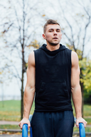 A portrait of a focused muscular young man in black workout clothes doing dips on parallel bars at...